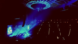 A concert venue illuminated with vibrant blue and purple lights, showcasing a large stage with performers. An overhead circular structure is present, adding to the modern ambiance. The audience is dimly visible, focusing their attention on the performance.