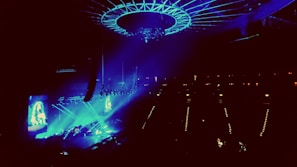 A concert venue illuminated with vibrant blue and purple lights, showcasing a large stage with performers. An overhead circular structure is present, adding to the modern ambiance. The audience is dimly visible, focusing their attention on the performance.