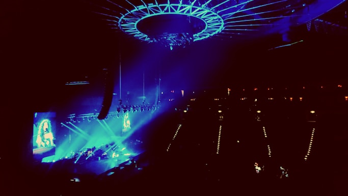 A concert venue illuminated with vibrant blue and purple lights, showcasing a large stage with performers. An overhead circular structure is present, adding to the modern ambiance. The audience is dimly visible, focusing their attention on the performance.