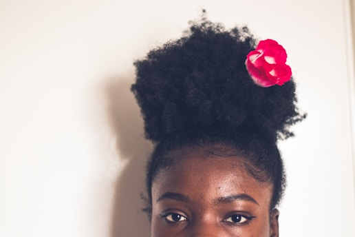 A person with natural, curly hair styled in a high afro puff adorned with a bright pink flower. The focus is on the top half of the face, showcasing expressive eyes with winged eyeliner.