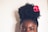 A person with natural, curly hair styled in a high afro puff adorned with a bright pink flower. The focus is on the top half of the face, showcasing expressive eyes with winged eyeliner.
