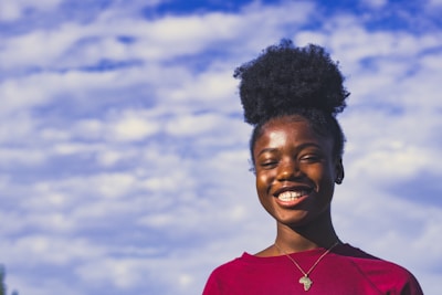 A person with a joyful expression, wearing a red shirt and an Africa-shaped pendant necklace, stands against a backdrop of a blue sky filled with fluffy white clouds. The natural light highlights their cheerful demeanor.