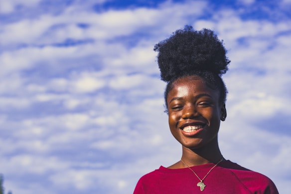A person with a joyful expression, wearing a red shirt and an Africa-shaped pendant necklace, stands against a backdrop of a blue sky filled with fluffy white clouds. The natural light highlights their cheerful demeanor.