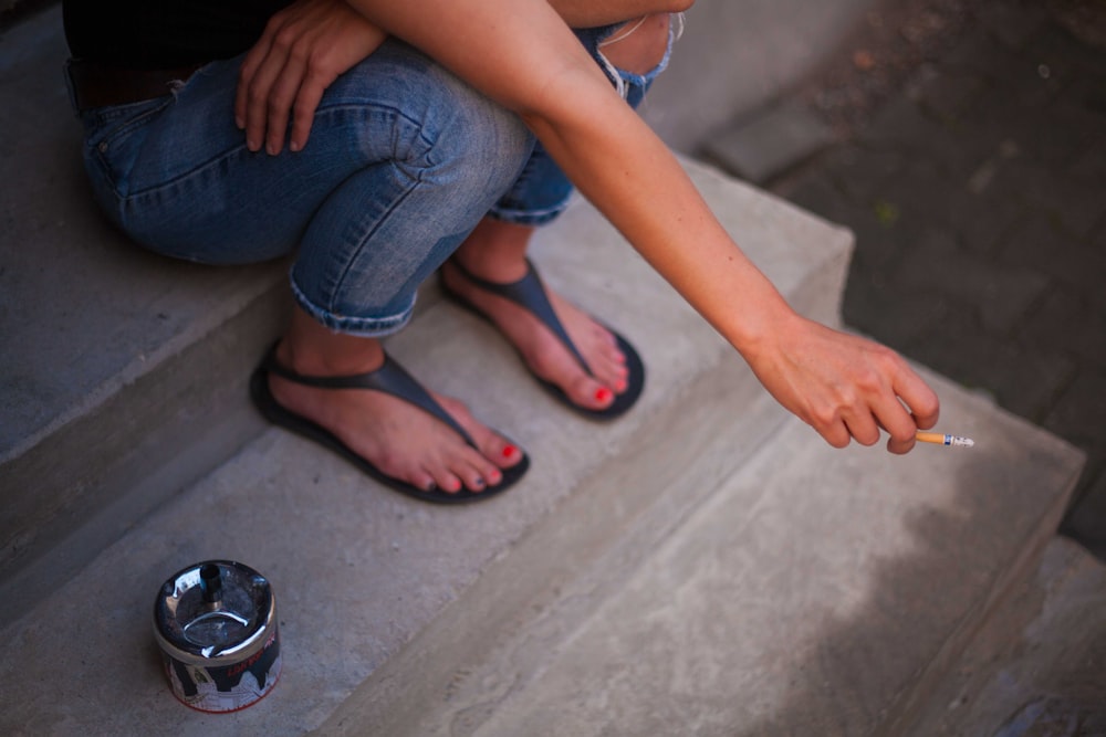 person wearing distressed blue denim jeans holding orange cigarette butt sitting on gray concrete stair