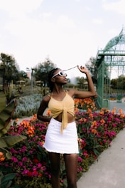 A fashionable individual stands among vibrant, colorful flowers and a pathway in a garden or park setting. They wear a yellow top and white skirt, accentuated by trendy sunglasses, with an elegant greenhouse structure in the background.
