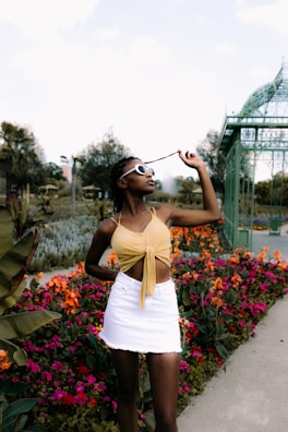 A fashionable individual stands among vibrant, colorful flowers and a pathway in a garden or park setting. They wear a yellow top and white skirt, accentuated by trendy sunglasses, with an elegant greenhouse structure in the background.