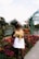 A fashionable individual stands among vibrant, colorful flowers and a pathway in a garden or park setting. They wear a yellow top and white skirt, accentuated by trendy sunglasses, with an elegant greenhouse structure in the background.