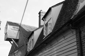 A vintage-style building with a steeply pitched roof featuring wooden siding and multiple gabled windows. An unusual structure juts out from the side, supported by beams. The architecture includes brick elements and overhanging eaves.