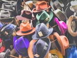 A collection of colorful hats displayed on stands, featuring a variety of styles including fedoras. Each hat has a price tag with handwritten numbers attached to the brim. The hats are in shades of orange, purple, red, green, and more, showcasing a vibrant assortment.