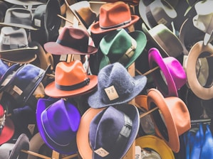 A collection of colorful hats displayed on stands, featuring a variety of styles including fedoras. Each hat has a price tag with handwritten numbers attached to the brim. The hats are in shades of orange, purple, red, green, and more, showcasing a vibrant assortment.