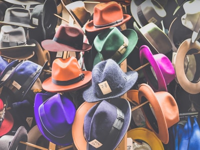 A collection of colorful hats displayed on stands, featuring a variety of styles including fedoras. Each hat has a price tag with handwritten numbers attached to the brim. The hats are in shades of orange, purple, red, green, and more, showcasing a vibrant assortment.