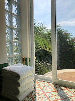 A stack of white towels is placed on a colorful tiled floor next to a large window. The window overlooks lush green palm trees and a clear blue sky. Glass blocks and green tiles add texture and color to the scene.