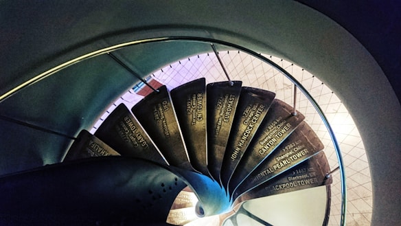 A spiraling staircase viewed from above, with each step displaying the names and distances to various famous towers around the world, such as the 'John Hancock Center' and 'Canton Tower'. The stairs are surrounded by a metal railing and descend in concentric circles, creating an optical illusion of depth.