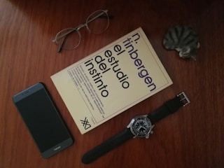 A book titled 'El estudio del instinto' is placed on a wooden surface alongside a pair of eyeglasses, a smartphone, a wristwatch, and a small cat figurine. The arrangement is neat and minimalistic.