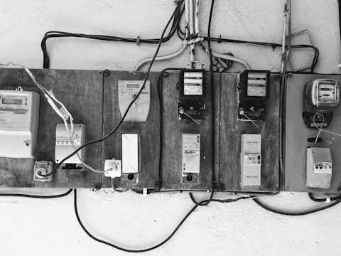 A row of old electric meters mounted on a wooden board are connected by various electrical wires. Some of the meters are encased, while others have exposed switches and dials. The setup appears rustic and utilitarian, with cables running horizontally and vertically.