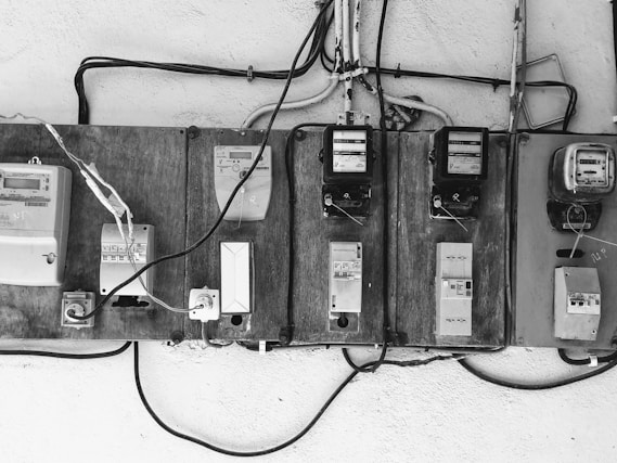 A row of old electric meters mounted on a wooden board are connected by various electrical wires. Some of the meters are encased, while others have exposed switches and dials. The setup appears rustic and utilitarian, with cables running horizontally and vertically.