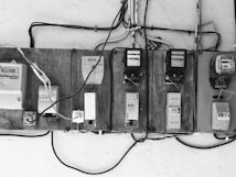 A row of old electric meters mounted on a wooden board are connected by various electrical wires. Some of the meters are encased, while others have exposed switches and dials. The setup appears rustic and utilitarian, with cables running horizontally and vertically.