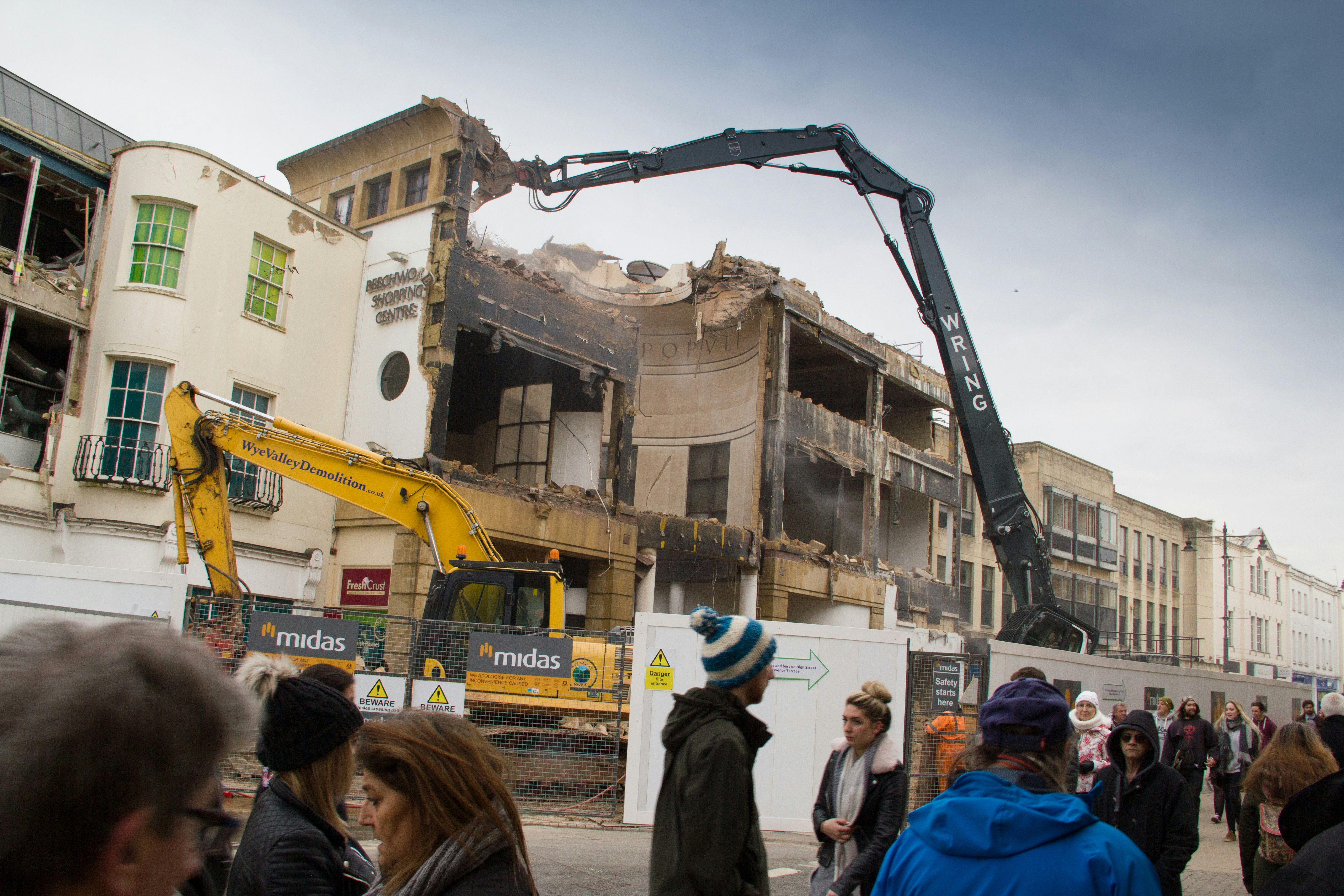 A partially demolished building is surrounded by construction equipment, including excavators. A crowd of people walks by, observing the demolition process. The building's facade is damaged, and rubble is visible. The scene takes place under a cloudy sky.
