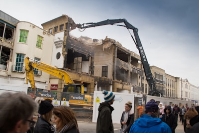 A partially demolished building is surrounded by construction equipment, including excavators. A crowd of people walks by, observing the demolition process. The building's facade is damaged, and rubble is visible. The scene takes place under a cloudy sky.