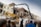 A partially demolished building is surrounded by construction equipment, including excavators. A crowd of people walks by, observing the demolition process. The building's facade is damaged, and rubble is visible. The scene takes place under a cloudy sky.