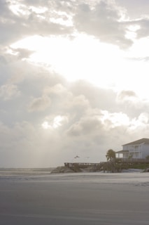A coastal scene featuring a sandy beach with a wooden pier extending out toward the ocean. A large house with a porch overlooks the beach, surrounded by palm trees. The sky is filled with dramatic clouds, and the sunlight creates a bright, glowing effect where it breaks through.