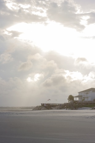 A coastal scene featuring a sandy beach with a wooden pier extending out toward the ocean. A large house with a porch overlooks the beach, surrounded by palm trees. The sky is filled with dramatic clouds, and the sunlight creates a bright, glowing effect where it breaks through.