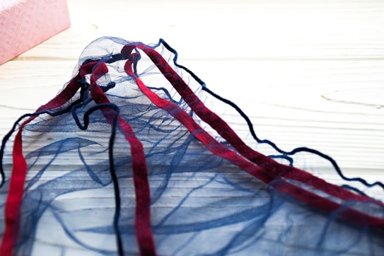 A piece of delicate blue mesh fabric with red and blue edges, resting on a light wooden surface. In the background is a pink textured box partially visible.