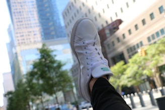 A white sneaker is prominently displayed in the foreground against an urban backdrop. The shoe is worn and visible from the side, with a green logo on the tongue. The background features tall, blurred buildings with glass facades and some greenery, likely trees, near the bottom right of the image.