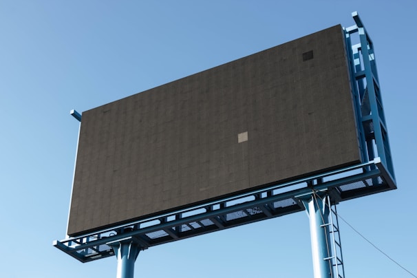 A large, empty billboard structure with a dark screen and blue frame stands against a clear blue sky. The billboard is elevated, supported by a metal framework with visible beams and pillars.