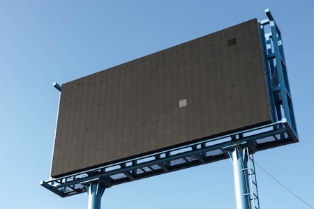 A large, empty billboard structure with a dark screen and blue frame stands against a clear blue sky. The billboard is elevated, supported by a metal framework with visible beams and pillars.