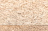 A textured surface composed of wood chips and strands oriented randomly and pressed together. The material appears natural and rustic, with variations in the shades of brown, beige, and tan.