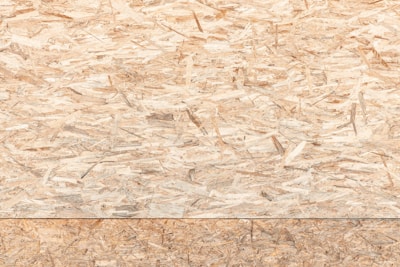 A textured surface composed of wood chips and strands oriented randomly and pressed together. The material appears natural and rustic, with variations in the shades of brown, beige, and tan.