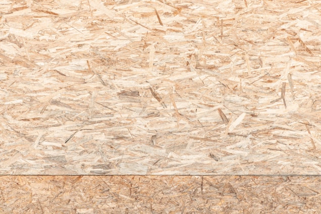 A textured surface composed of wood chips and strands oriented randomly and pressed together. The material appears natural and rustic, with variations in the shades of brown, beige, and tan.