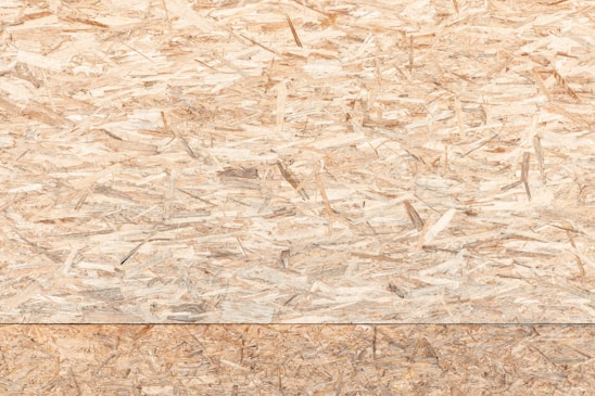 A textured surface composed of wood chips and strands oriented randomly and pressed together. The material appears natural and rustic, with variations in the shades of brown, beige, and tan.