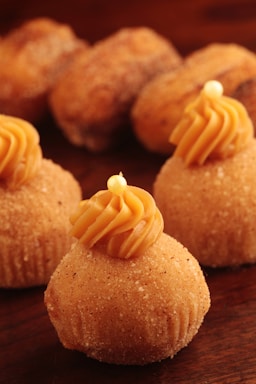 Several sugar-coated pastries are topped with a swirl of caramel-colored frosting, each adorned with a small decorative pearl. The pastries are arranged on a dark wooden surface, creating a warm and inviting atmosphere.