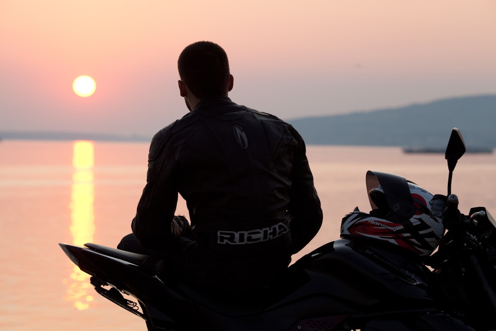 silhouette photo of man sitting on sports bike beside sea during golden hour