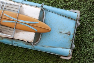 A vintage toy model of a blue car with a surfboard attached to its roof, placed on a grassy surface. The car and surfboard appear worn, with visible rust and weathering, giving it a nostalgic appearance.