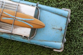 A vintage toy model of a blue car with a surfboard attached to its roof, placed on a grassy surface. The car and surfboard appear worn, with visible rust and weathering, giving it a nostalgic appearance.