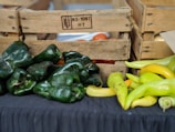 A collection of green and yellow peppers placed on a black cloth surface. Wooden crates with inscriptions are stacked in the background, containing tomatoes and possibly other vegetables.