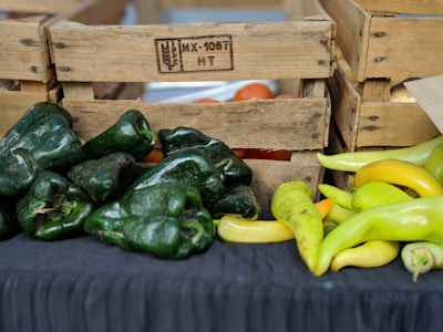 A collection of green and yellow peppers placed on a black cloth surface. Wooden crates with inscriptions are stacked in the background, containing tomatoes and possibly other vegetables.