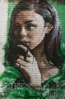 A mural on a brick wall depicts a woman's face with long hair, holding a strand of it near her mouth. The artwork is detailed, showcasing realistic shadows and highlights that give depth to the woman's expression. The background is shaded with hues of green, adding contrast to her dark hair and soft features.