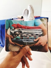 A person holding a book with an image of hands holding a stack of colorful folded clothes. The background shows a pair of white sneakers on someone’s feet resting on a table, with a blue draped fabric and rolls of paper towels visible.