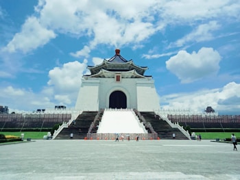 A grand, traditional Chinese-style building with a striking blue and white color scheme towers against a backdrop of a bright blue sky dotted with fluffy clouds. The structure features a large staircase leading up to an expansive entrance adorned with a wooden sign. The foreground shows an open plaza with a few people walking, surrounded by green lawns.
