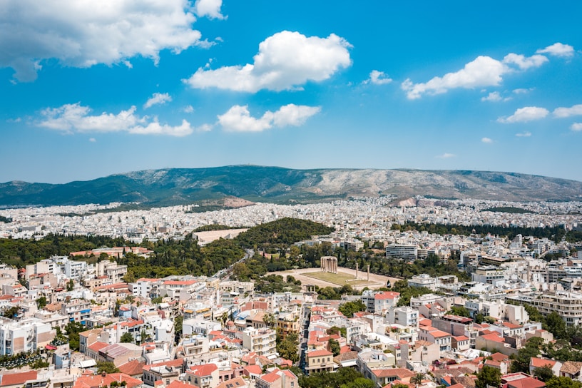 An expansive view of a cityscape with clusters of white and beige buildings. In the foreground, there are red-roofed structures, and in the center, an ancient ruin with tall columns surrounded by greenery. Beyond the city, a range of mountains stretches across the horizon under a partly cloudy blue sky.