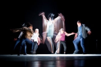The image captures a dynamic dance performance with several figures in motion, creating a blur effect that emphasizes movement and energy against a dark background.