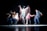 The image captures a dynamic dance performance with several figures in motion, creating a blur effect that emphasizes movement and energy against a dark background.