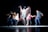 The image captures a dynamic dance performance with several figures in motion, creating a blur effect that emphasizes movement and energy against a dark background.