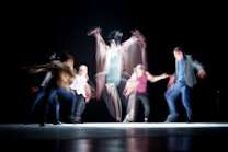 The image captures a dynamic dance performance with several figures in motion, creating a blur effect that emphasizes movement and energy against a dark background.