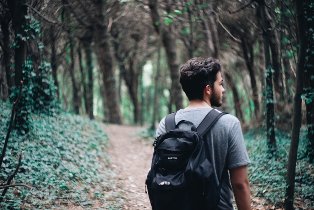 Mann in grauem T-Shirt und schwarzem Rucksack, der tagsüber auf dem Weg zwischen Bäumen spazieren geht