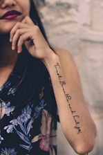 A woman with a tattoo on her forearm featuring cursive writing. She is wearing a dark floral-patterned garment and has dark lipstick. Her hand is near her face, and the background is a light-colored textured wall.